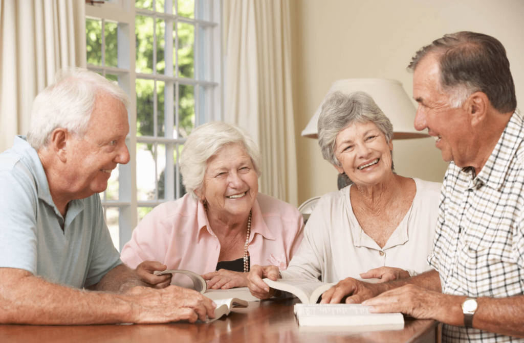 A group of seniors gathered around a table talking, smiling and reading a book.